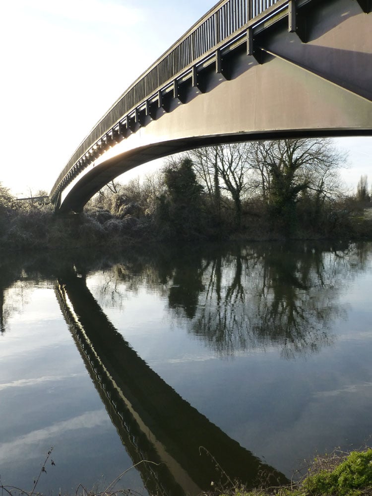 Footbridge over the Thames at Bray, Berkshire, with mirror reflection of arch in the still waters of the river and sun glinting off the structure of the bridge